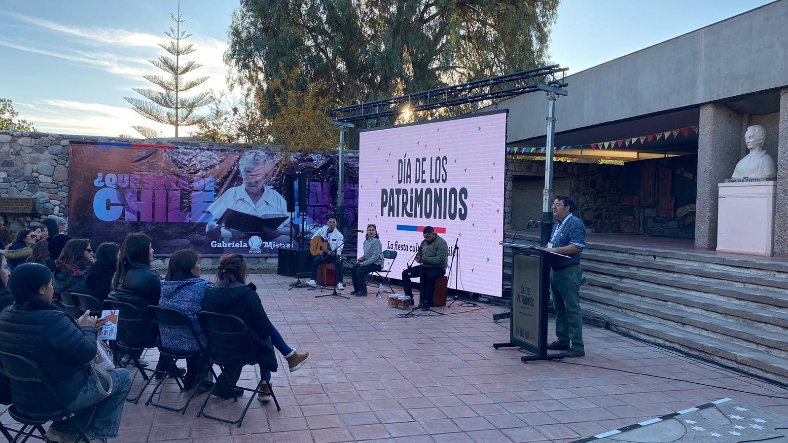 Frontis del museo con una pantalla gigante en la que se lee "Día de los Patrimonios", un trío de músicos a punto de tocar y el director del museo entregando un discurso a un público de unas 40 personas sentadas en sillas plegables.