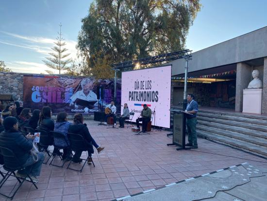 Frontis del museo con una pantalla gigante en la que se lee "Día de los Patrimonios", un trío de músicos a punto de tocar y el director del museo entregando un discurso a un público de unas 40 personas sentadas en sillas plegables.